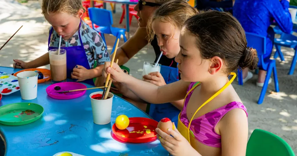 Children painting crafts at a blue outdoor table with brushes and colorful paints